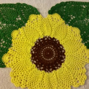 Puede incluir: Un encaje de ganchillo de girasol con hojas verdes. El girasol es amarillo con un centro marrón. El encaje está hecho con un punto de ganchillo delicado.
