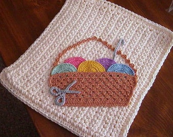 May include: A white crocheted square with a brown crocheted basket filled with colorful yarn balls. The basket has a crocheted handle and a pair of crocheted scissors.
