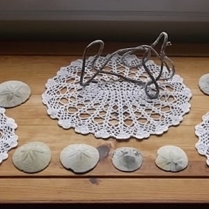 May include: Four white crocheted doilies with a decorative metal sculpture in the center. Each doily has a sand dollar on top. The doilies are on a wooden table.