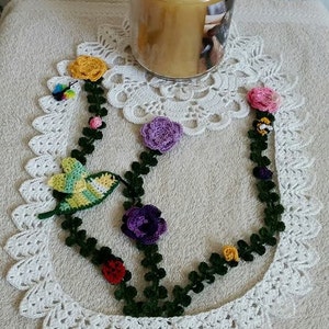 Pode incluir: Um crochê branco com uma guirlanda de crochê verde decorada com flores e pássaros de crochê coloridos. A guirlanda é adornada com pequenas abelhas de crochê. Um frasco de vidro com tampa cheio de uma substância amarela está no fundo.