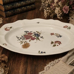 Peut inclure: Un plat de service ovale en céramique blanche avec un bord festonné et un motif floral central composé de fleurs colorées dans des tons de rouge, bleu et jaune, avec de petites touches florales supplémentaires dispersées sur le bord.