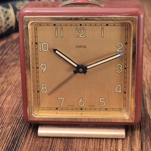 May include: A square Oris alarm clock with a maroon casing and a gold-toned face. The clock features black numerals, gold-colored hands, and a small red accent on the top button. The text "ORIS" is printed in the center, with "SWISS MADE" at the bottom.