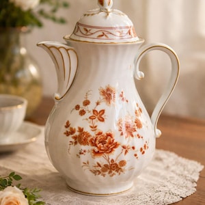 May include: White ceramic coffee pot with a lid, adorned with an orange and brown floral pattern. The pot has a curved handle and gold trim. A decorative piece for serving beverages.