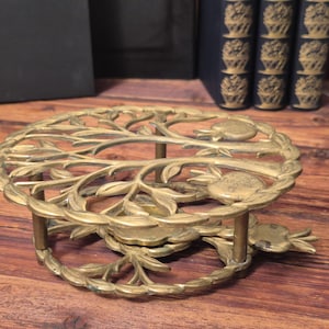 Könnte beinhalten: Ein Vintage-Topfuntersetzer aus Messing mit einem kreisförmigen Design mit einem Baum- und Fruchtmotiv. Der Topfuntersetzer hat ein erhabenes Design und wird von kurzen Beinen getragen. Das Messing hat eine warme, gealterte Patina.