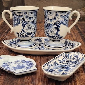 Könnte beinhalten: Zwei weiße Keramikbecher mit blauen Blumenmustern auf einem passenden ovalen Tablett, begleitet von einem kleinen Teebeutelhalter mit der Aufschrift TEA BAG und einer rechteckigen Schale, alle mit einheitlichen blau-weißen Blumenmotiven.