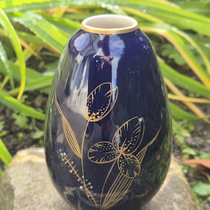 May include: A navy blue ceramic vase with a gold rim and floral design. The vase has a glossy finish and a rounded shape. Gold-coloured floral patterns adorn the front of the vase, adding a touch of elegance.