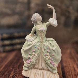 Può includere: Una statuetta in porcellana raffigurante una persona con un abito verde pallido e una sottogonna color crema. L'abito presenta applicazioni floreali rosa e blu, un corpetto rifinito in pizzo e maniche a balze. Un braccio è sollevato verso l'alto.