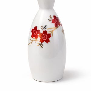 Puede incluir: Botella de sake de cerámica blanca con borde dorado y diseño floral rojo. La botella tiene un cuello delgado y un cuerpo redondeado. Las flores rojas están acentuadas con tallos y hojas doradas.