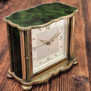 May include: A rectangular mantel clock with a dark green marbled top and base, featuring a brass frame and four small feet. The white clock face has a central grid pattern, gold-toned hands, and the text Jaz and Made in France printed on it.