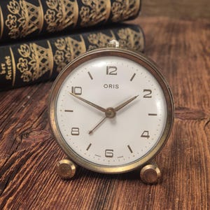 May include: A round Oris alarm clock with a white face, gold-toned metal casing, and two small circular feet. The clock features black Arabic numerals at the 12, 2, 4, 6, 8, and 10 positions, with thin gold hands and the text Oris Swiss Made.
