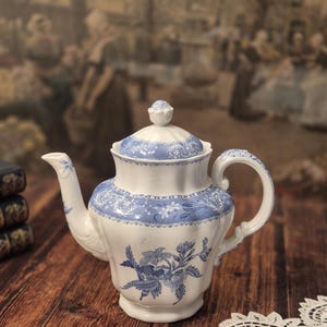 May include: A white porcelain teapot featuring a blue floral pattern and a matching lid with a finial. The teapot has a curved handle, a spout, and a scalloped rim design. The body is decorated with intricate blue botanical illustrations.