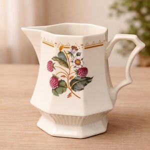 Op de afbeelding: Crèmekleurige keramische kan met een decoratief frambozen- en bloemmotief. De kan heeft een uniek handvat en een geometrische basis. Gouden randen accentueren de bovenrand, wat een vleugje elegantie toevoegt aan dit keukengerei.