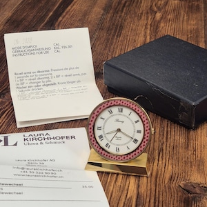 Può includere: Una sveglia analogica rotonda con quadrante bianco, numeri romani neri e un bordo a pois rosa e bianchi. L'orologio è montato su una base trapezoidale dorata ed è accompagnato da una scatola nera e da fogli di istruzioni stampati.