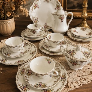 Puede incluir: Un juego de té de porcelana blanca con un colorido estampado floral y bordes dorados. El conjunto incluye varias tazas, platillos, platos, una tetera con tapa y un azucarero, dispuestos sobre una superficie con detalles de encaje.