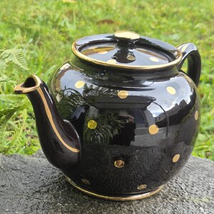Può includere: Una teiera in ceramica nera con pois dorati e finiture dorate. La teiera ha una forma arrotondata, un beccuccio curvo e un manico. Il coperchio ha anche un pomello dorato. La teiera è appoggiata su una superficie di pietra.