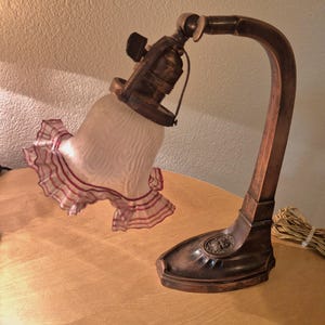 Puede incluir: Una lámpara de escritorio antigua con un brazo y una base curvados de color bronce. La lámpara presenta una pantalla de cristal esmerilado con un borde con volantes en blanco y rojo. La lámpara está sobre una superficie de madera.