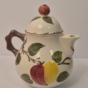 May include: Cream-colored ceramic teapot with a lid and a brown handle. The teapot is decorated with green leaves and a split apple design in red and yellow. The lid is topped with a small, brown apple-shaped knob.