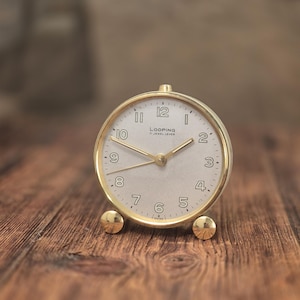 May include: A round gold-tone alarm clock with a light-colored face, featuring the text "LOOPING" and "15 JEWEL LEVER" printed in black. The clock has gold-tone hands, Arabic numerals, and two small gold-tone spherical feet at the base.