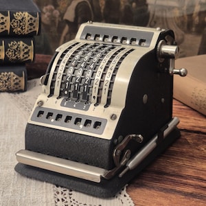 Könnte beinhalten: Ein mechanischer Vintage-Taschenrechner mit schwarzem, strukturiertem Gehäuse und cremefarbener Metallfront. Er verfügt über vertikale Schieberegler für Zahlen, eine Reihe von numerischen Anzeigefenstern, eine seitliche Metallkurbel und einen Stift.