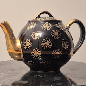 May include: A black ceramic teapot with a gold-colored spout, handle, and lid handle. The teapot is decorated with a pattern of gold-colored daisy flowers. The teapot is sitting on a dark surface.