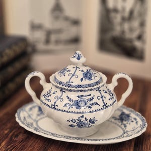 Op de afbeelding: Een witte keramische suikerpot met deksel en twee oren, voorzien van een blauw bloemmotief en een kleine vogelillustratie. De pot staat op een bijpassende ovale schotel, beide gedecoreerd met blauwe ranken en geschulpte randen.
