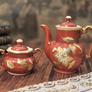 Può includere: Una teiera in ceramica color terracotta e una zuccheriera abbinata con coperchio. Entrambi i pezzi presentano un bordo color crema, un manico rifinito in oro e un motivo floreale dipinto con foglie bianche, dorate e verdi sulla superficie.