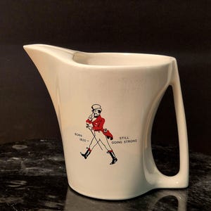 May include: Cream-colored ceramic pitcher with a curved handle and spout. The pitcher features a graphic of a man in a red coat and the text "BORN 1820 - STILL GOING STRONG". The pitcher is on a dark, patterned surface.