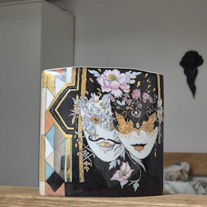 Può includere: Vaso rettangolare in ceramica nera con un design decorativo raffigurante due maschere veneziane. Le maschere sono bianche e dorate, ornate da accenti floreali. Il vaso presenta un motivo dorato e geometrico sul lato.