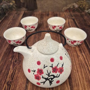 Puede incluir: Un juego de té de cerámica que consta de una tetera redonda con asa de metal negro y cuatro tazas pequeñas a juego. Las piezas son de color blanco roto con textura moteada, decoradas con ramas negras y flores de ciruelo de color rojo.
