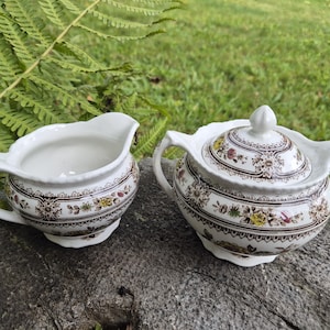 Könnte beinhalten: Ein weißes Keramik-Sahnekännchen und eine Zuckerdose mit Deckel. Die Teile haben ein braunes Blumenmuster mit gelben und roten Akzenten. Die Zuckerdose hat einen Deckel mit einem kleinen Knauf.