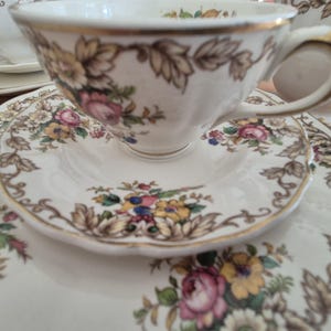 May include: A vintage tea set comprising a teacup, saucer, and plate. The white porcelain items are decorated with a floral pattern in pink, yellow, and blue, with brown leaves and gold edging. The teacup sits on the saucer, with a matching plate beneath.