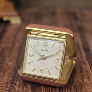 May include: A square Europa travel alarm clock with a tan leather-like case. The clock face is white with gold-toned hands and hour markers, featuring the text Europa and 2 Jewels. The clock is hinged and open, revealing a gold-toned metal frame.