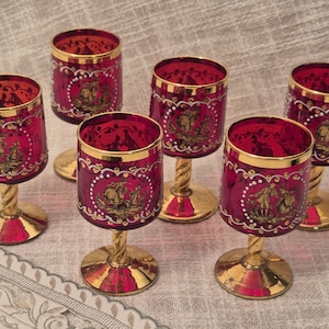 May include: A set of five ruby red glass goblets featuring gold-rimmed edges and gold-toned twisted stems. Each glass displays a white circular frame containing a detailed gold-colored illustration of figures in a landscape setting.