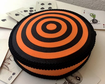 Puede incluir: Una caja redonda de color naranja y negro en forma de diana con un borde negro. La caja está sentada sobre una superficie blanca con cartas de juego alrededor.
