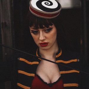 May include: A woman wearing a black and gold striped jacket with a red top and a black and white spiral hat. She is holding a black whip.
