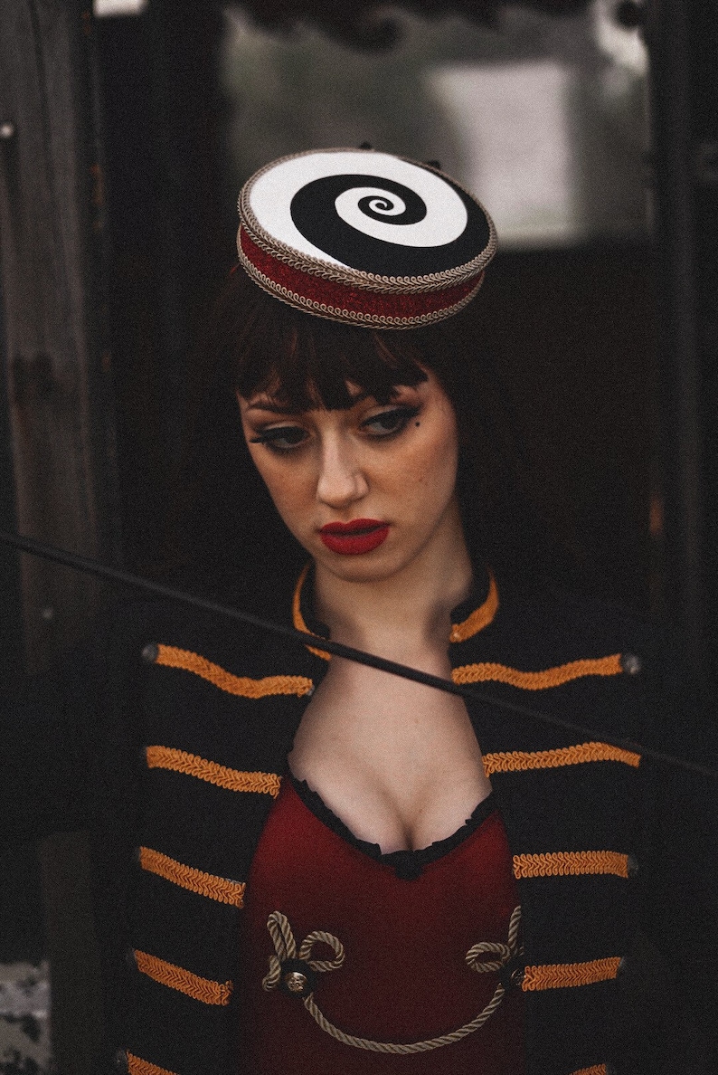 May include: A woman wearing a black and gold striped jacket with a red top and a black and white spiral hat. She is holding a black whip.