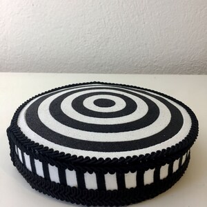 Puede incluir: Un soporte para tartas redondo, blanco y negro, con un dise&ntilde;o de c&iacute;rculos conc&eacute;ntricos. El soporte est&aacute; decorado con una cinta a rayas blancas y negras y un ribete trenzado negro.