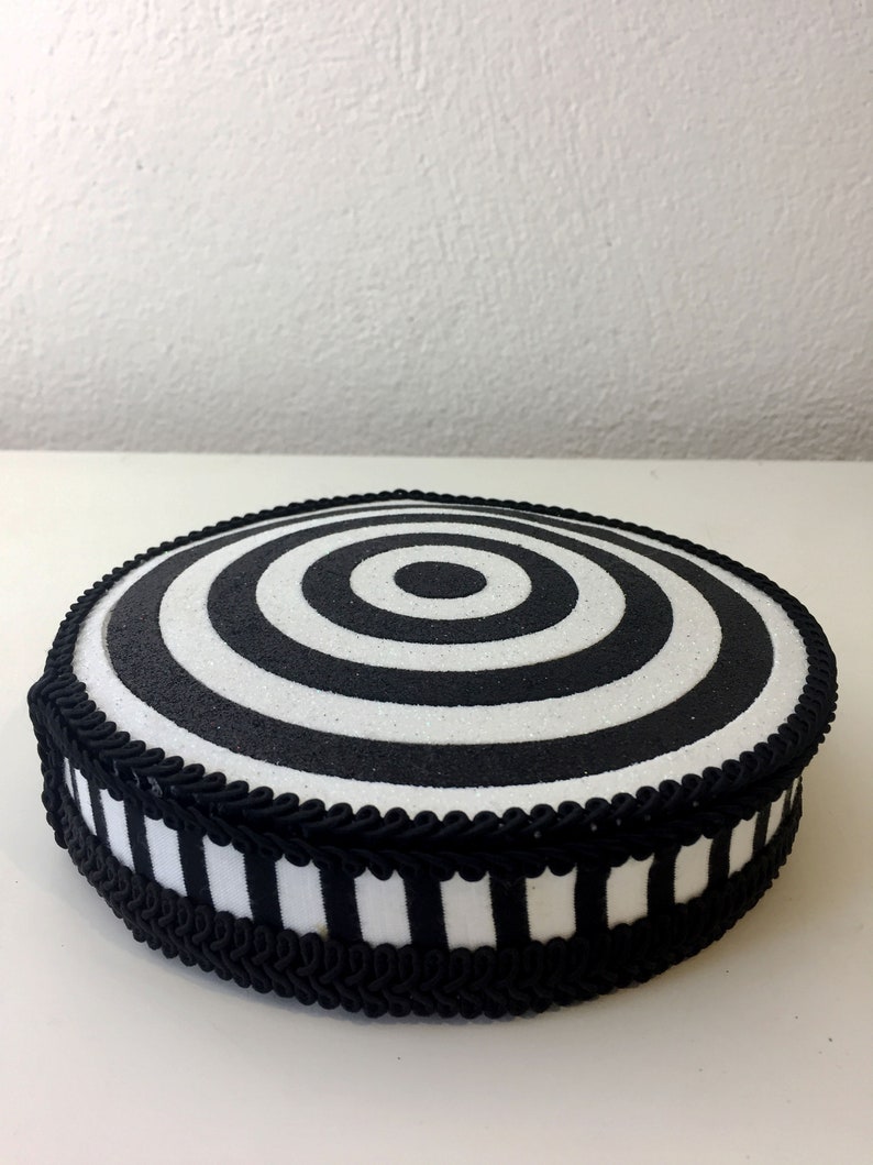 Puede incluir: Un soporte para tartas redondo, blanco y negro, con un dise&ntilde;o de c&iacute;rculos conc&eacute;ntricos. El soporte est&aacute; decorado con una cinta a rayas blancas y negras y un ribete trenzado negro.