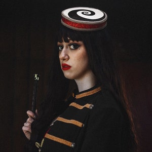 May include: A woman wearing a black and white spiral patterned hat with a red band, a black jacket with gold trim, and red lipstick. She is holding a black stick in her right hand.