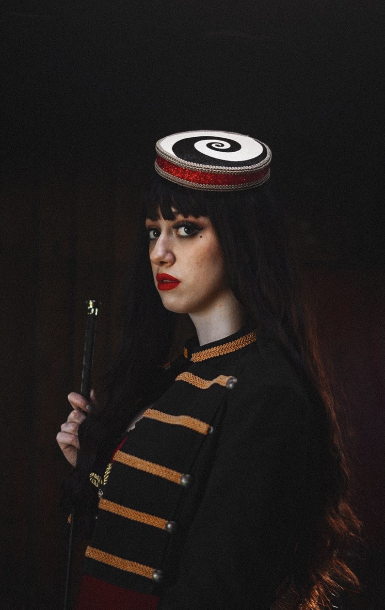 May include: A woman wearing a black and white spiral patterned hat with a red band, a black jacket with gold trim, and red lipstick. She is holding a black stick in her right hand.