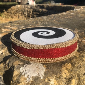 May include: A round, black and white spiral design with a red glitter trim and gold braid around the edge. The design is on a round, white base.