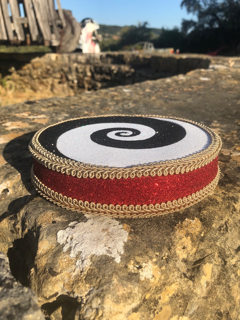 May include: A round, black and white spiral design with a red glitter trim and gold braid around the edge. The design is on a round, white base.