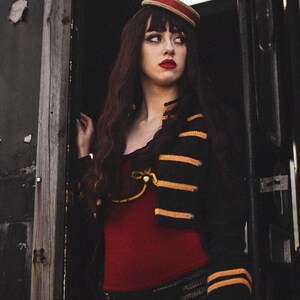 May include: A woman wearing a black and gold striped jacket with red trim, a red top, black shorts, and fishnet stockings. She is wearing a red and white hat and has dark hair.