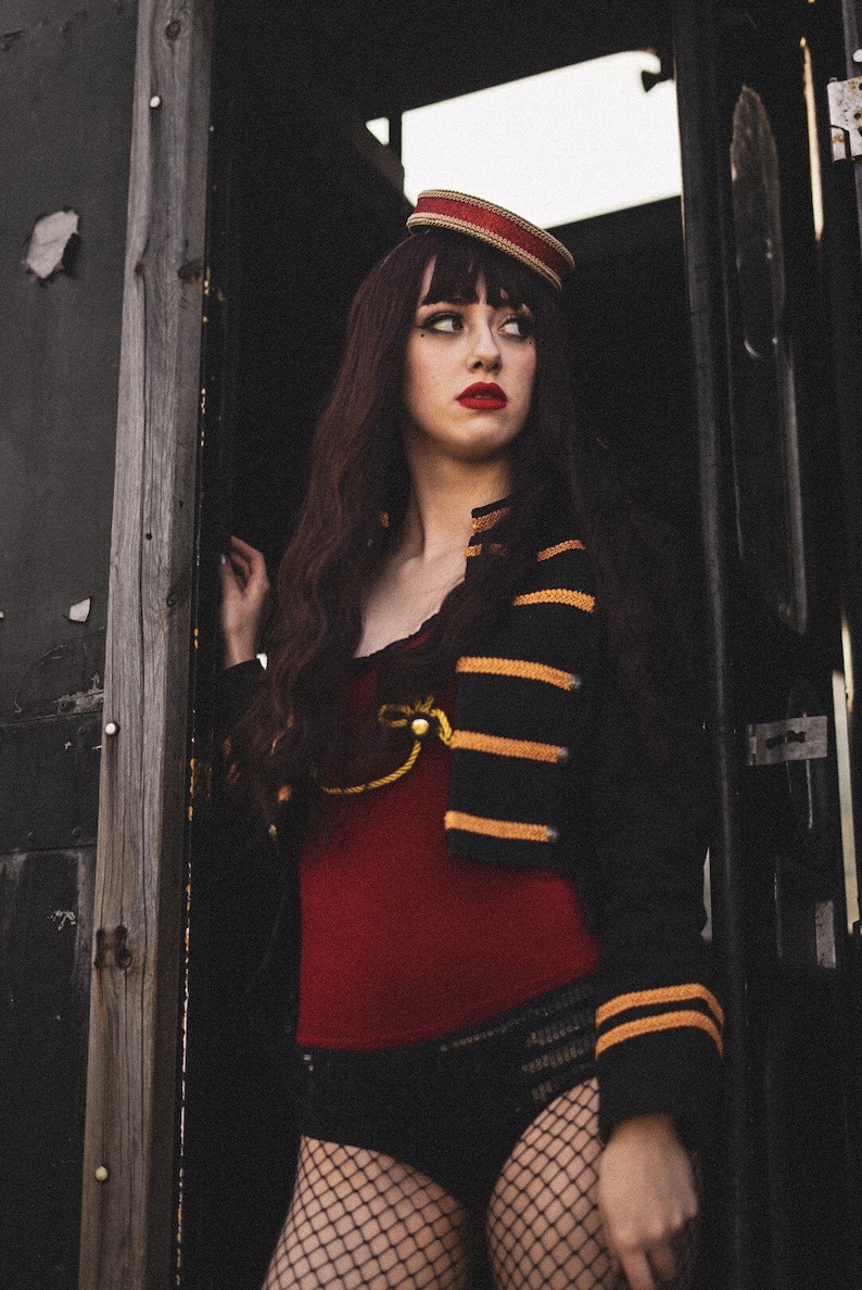 May include: A woman wearing a black and gold striped jacket with red trim, a red top, black shorts, and fishnet stockings. She is wearing a red and white hat and has dark hair.