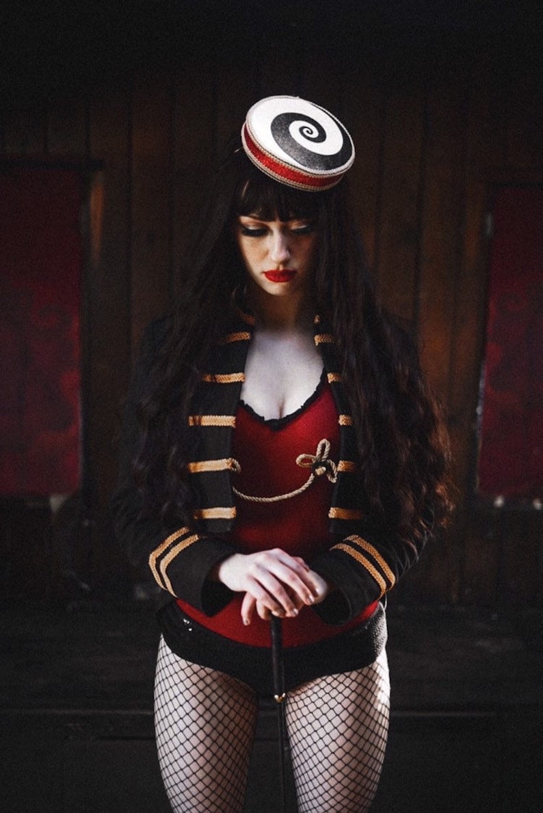 May include: A woman wearing a black and gold jacket with red trim, a red bodysuit, and black fishnet stockings. She is holding a black cane and wearing a white and black spiral hat.