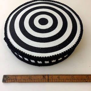 Puede incluir: Objetivo circular blanco y negro con una superficie brillante y un borde trenzado negro alrededor del borde. El objetivo tiene aproximadamente 12,5 cm de di&aacute;metro.