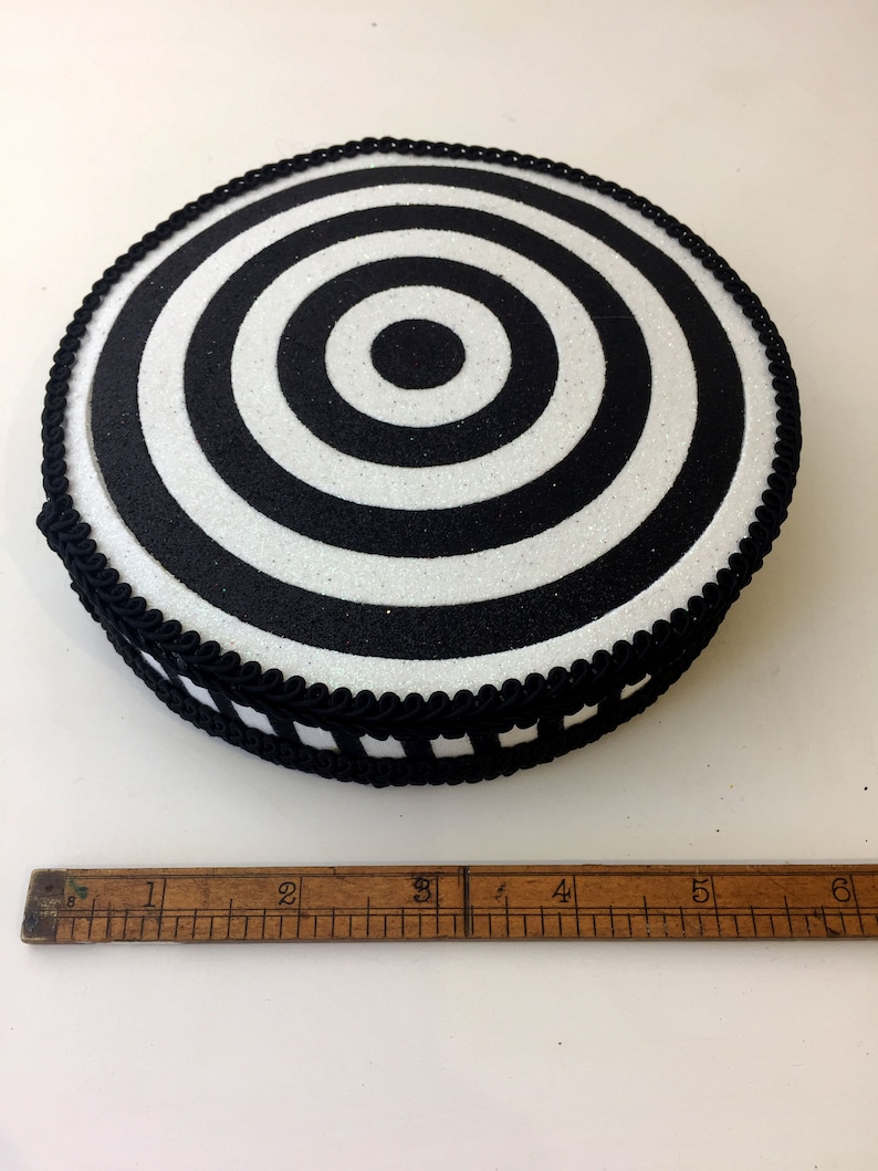 Puede incluir: Objetivo circular blanco y negro con una superficie brillante y un borde trenzado negro alrededor del borde. El objetivo tiene aproximadamente 12,5 cm de di&aacute;metro.