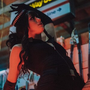 Puede incluir: Una mujer con un atuendo negro, un sombrero a rayas blanco y negro, medias de red y guantes negros. El fondo es una pared roja y azul con un letrero amarillo que dice "Way Out" con una flecha.
