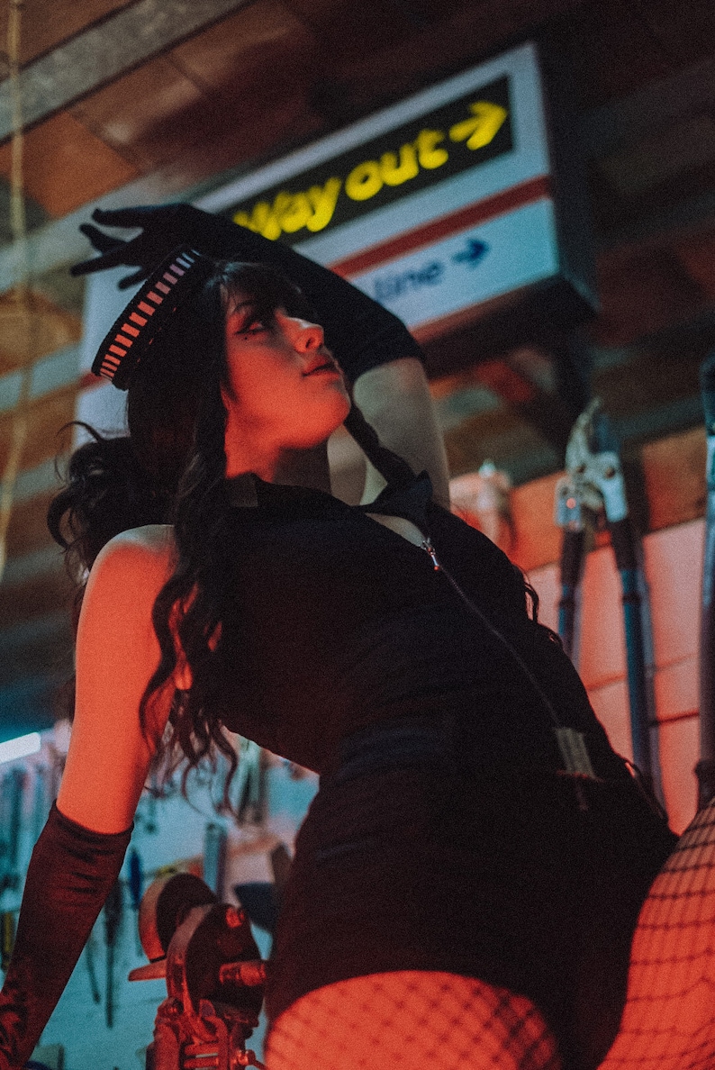Puede incluir: Una mujer con un atuendo negro, un sombrero a rayas blanco y negro, medias de red y guantes negros. El fondo es una pared roja y azul con un letrero amarillo que dice "Way Out" con una flecha.