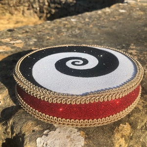 May include: A round cake topper with a black and white spiral design. The topper is decorated with gold trim and a red glitter ribbon.